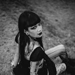 A black and white portrait of a woman with tattoos and gothic style, sitting outdoors on grass.